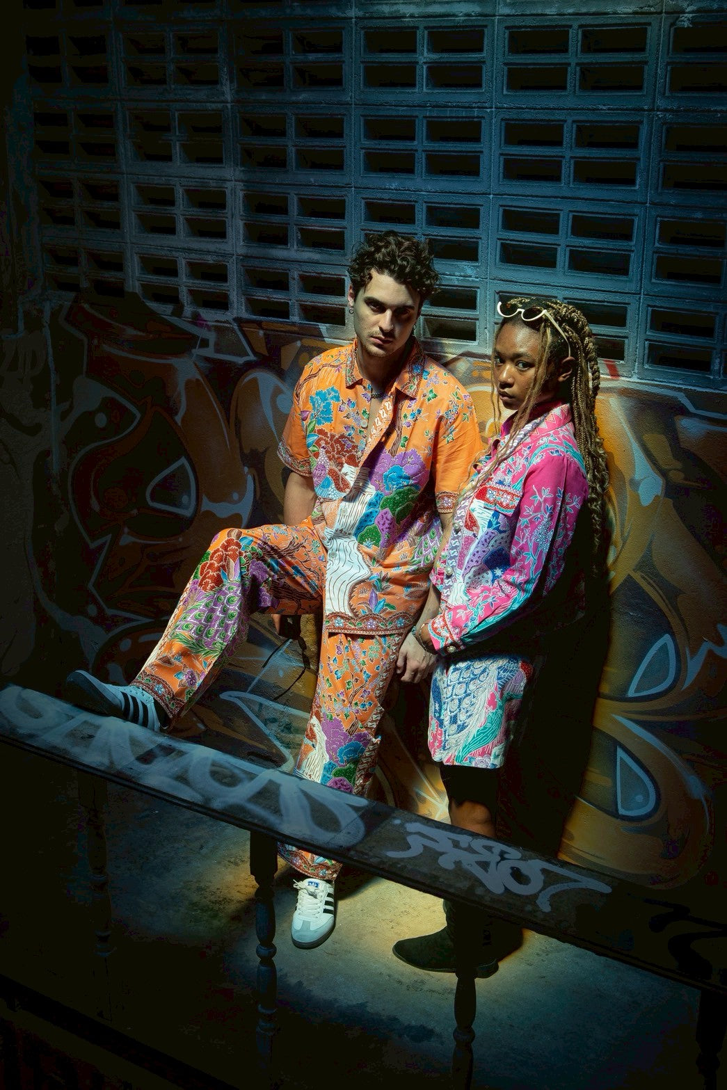 Two people in colorful outfits sitting on a bench with graffiti in the background